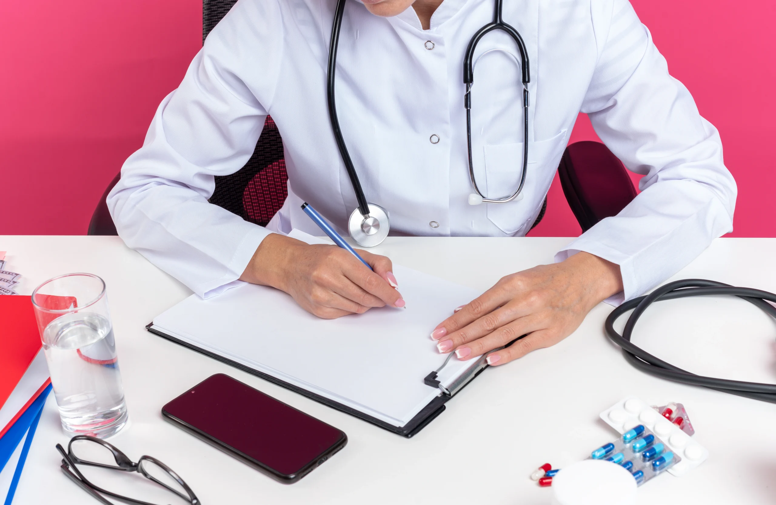 pleased-adult-slavic-female-doctor-medical-robe-with-stethoscope-sitting-desk-with-office-tools-writing-clipboard-isolated-pink-wall-with-copy-space